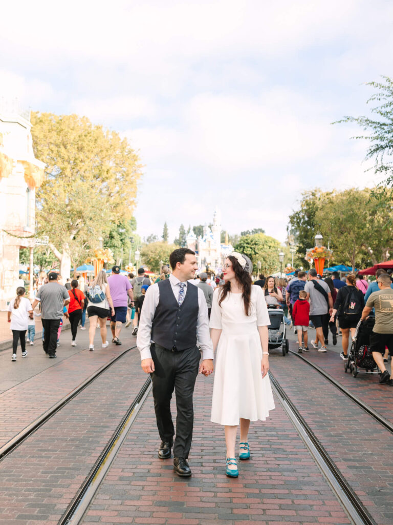 A couple in formal attire walks hand in hand down a bustling Disneyland theme park street, smiling at each other.