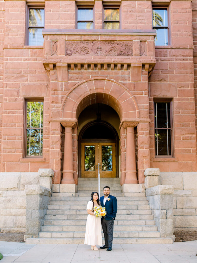 A couple stands on stone steps in front of a historic red brick building with an arched entrance.
