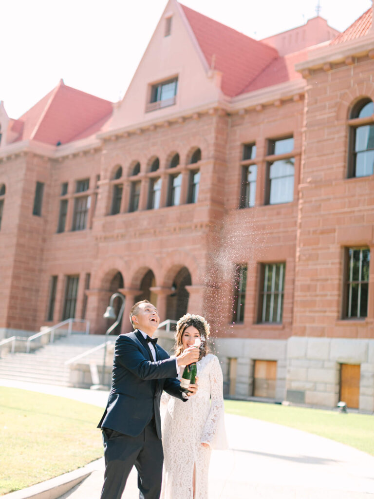 A couple in formal attire joyfully pops a champagne bottle, with fizzy spray in the air.