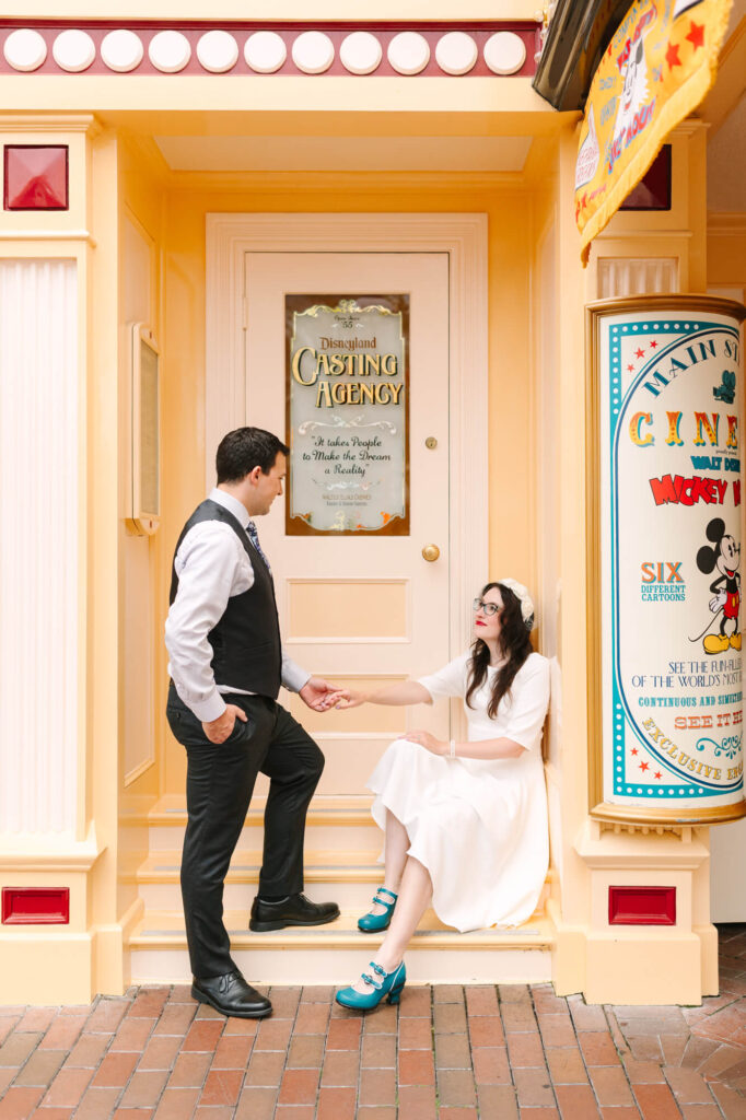 A man in a formal vest and tie holds hands with a woman in a white dress sitting on a step. They are in front of a vintage-style door labeled "Disneyland Casting Agency" with a colorful Mickey Mouse poster nearby.
