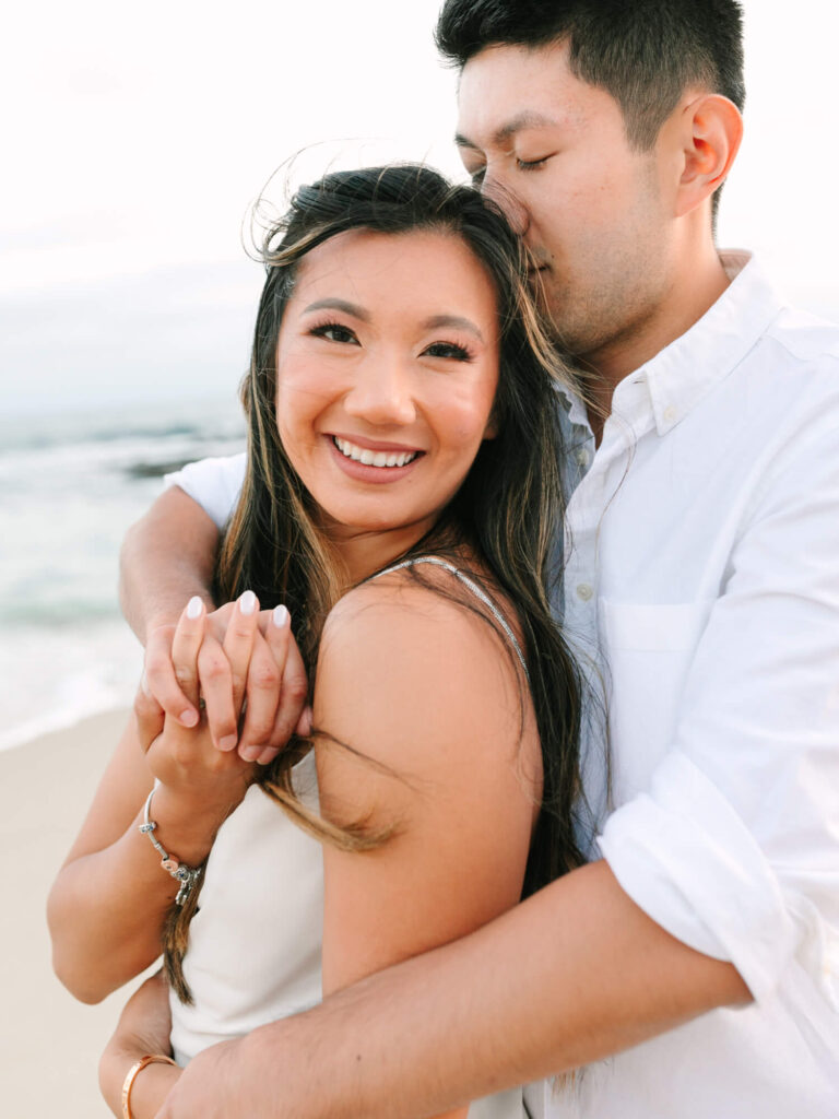 A couple embraces on a beach at sunset. The woman smiles brightly, and the man gently kisses her head.