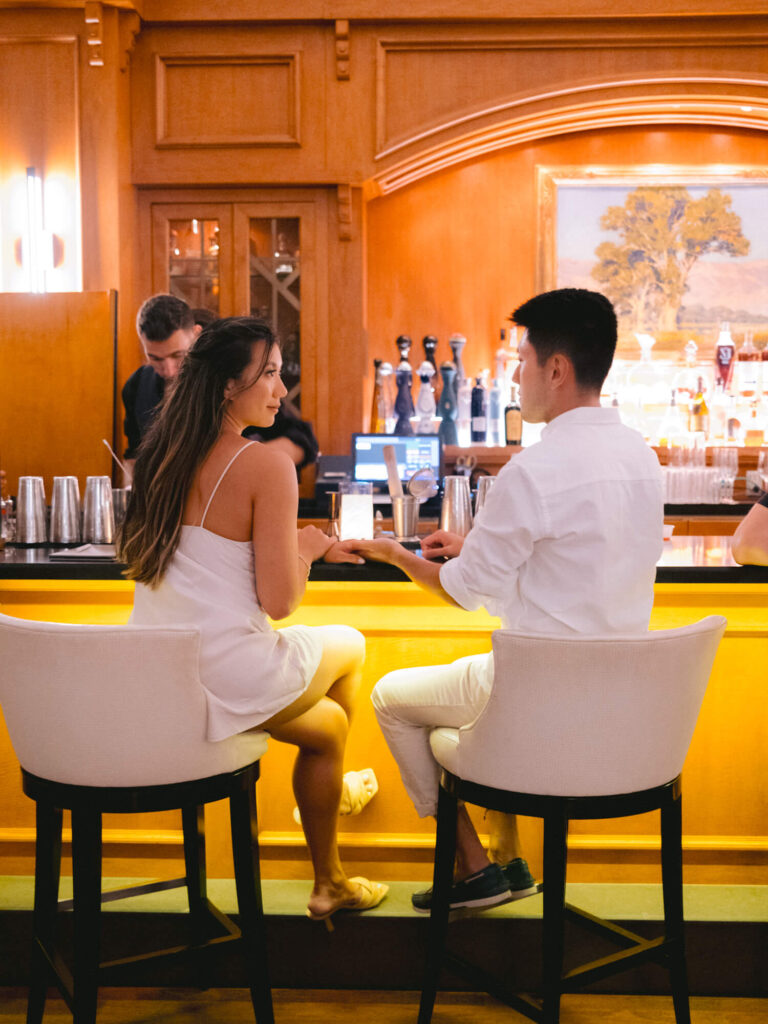 A couple in white clothes sits at a warmly lit bar, engaged in a lively conversation, with a bartender preparing drinks in the background.