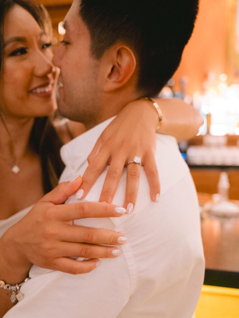 A couple embracing, smiling warmly at each other. The woman shows an engagement ring on her finger.