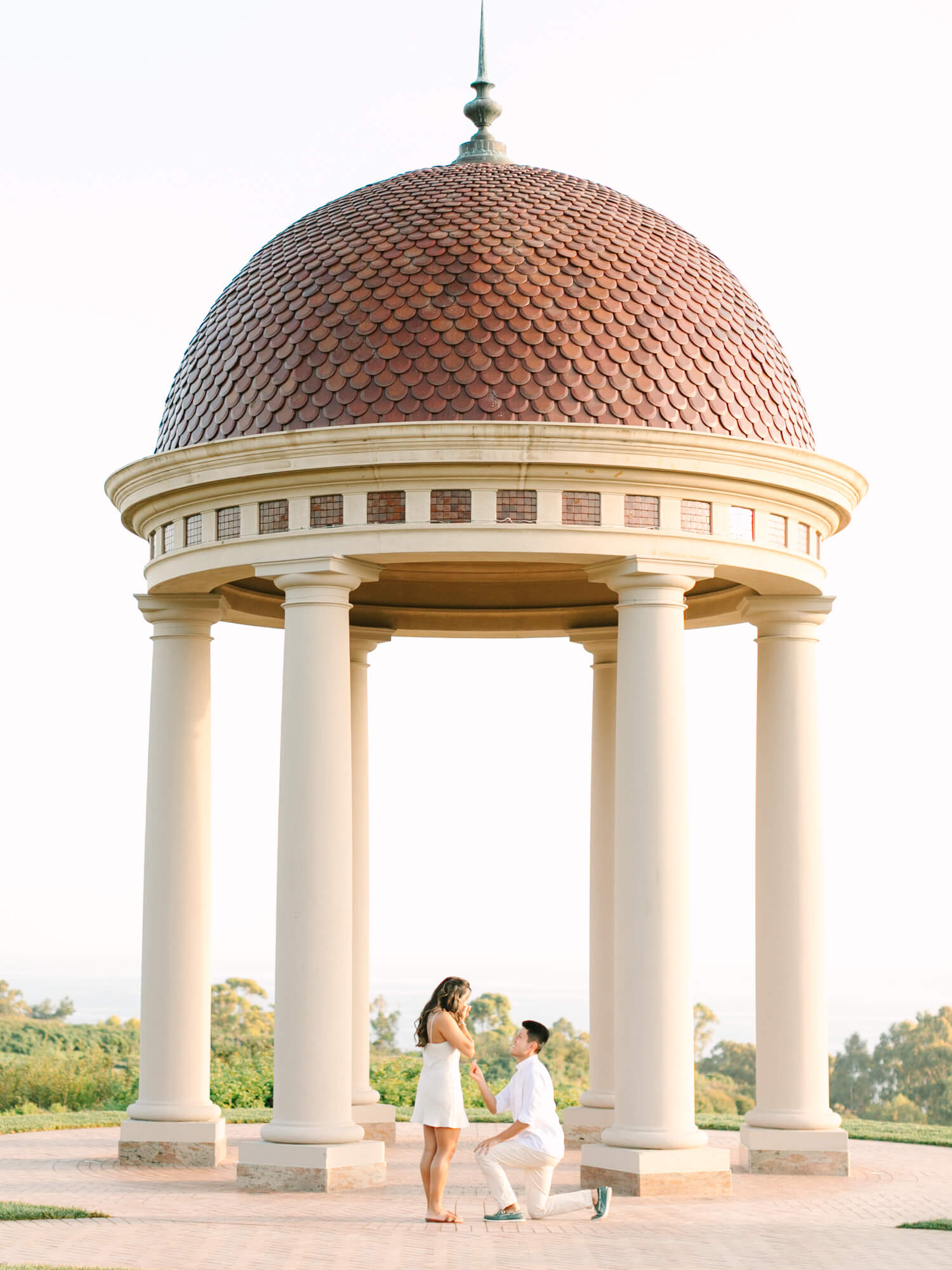 A man kneels in front of a woman, proposing under a domed gazebo with tall columns.