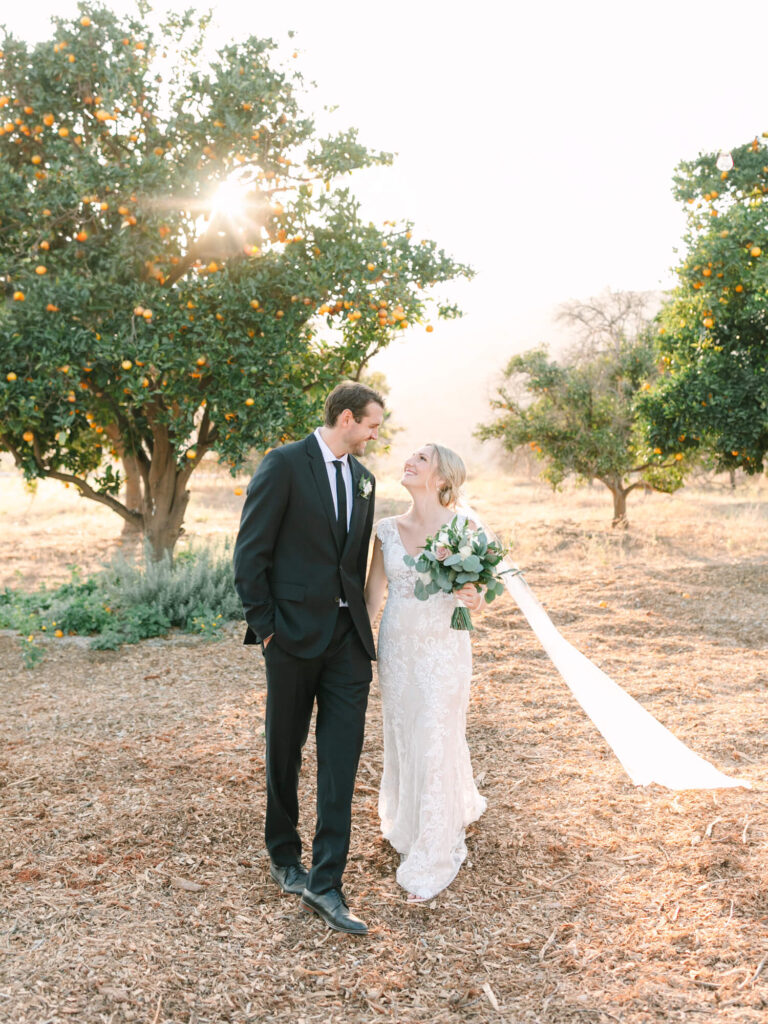 Bride and groom walking under orange trees, sunlight filtering through leaves.