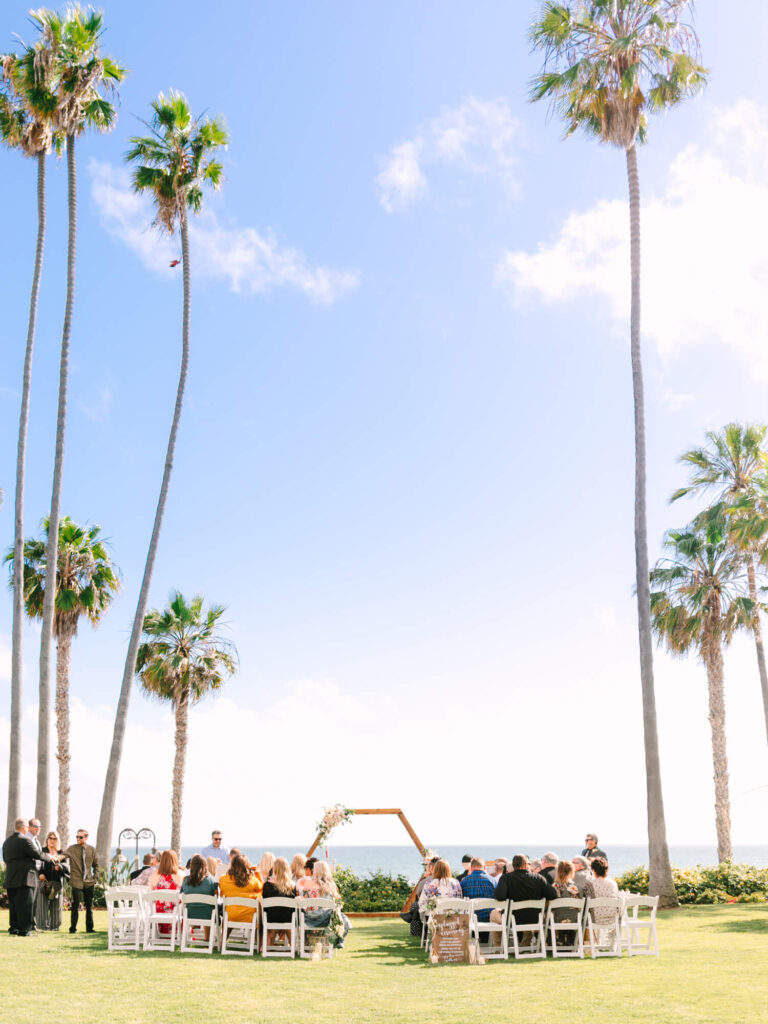 Outdoor wedding ceremony by the ocean with tall palm trees, clear blue sky, and hexagonal wooden arch.
