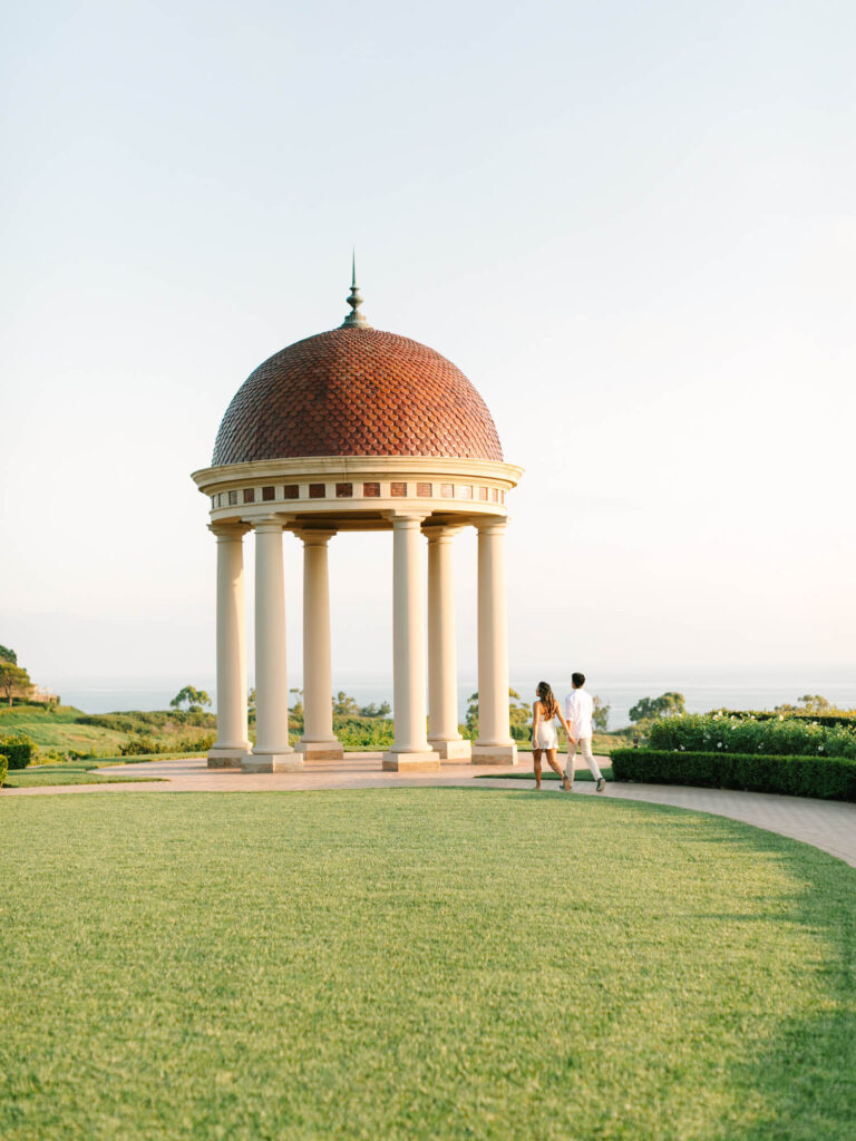 A couple walks towards a domed gazebo with columns, set in a lush green lawn against a backdrop of blue sky and ocean.