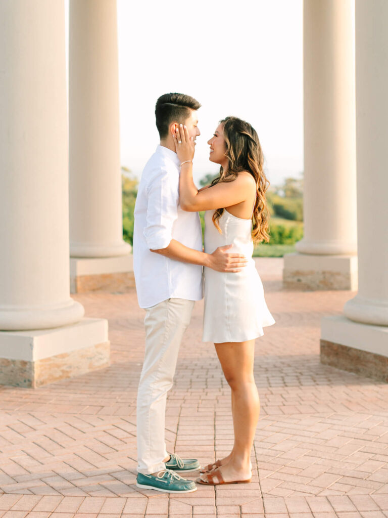 A couple stands in front of large columns, gazing at each other lovingly. The woman in a white dress touches the man's face.