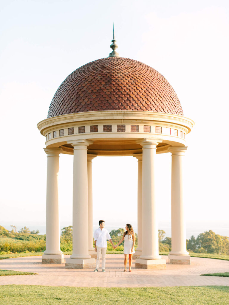 A couple in white clothes holds hands under a domed gazebo with tall columns.