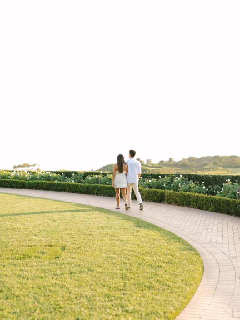 A couple walks hand in hand on a paved path surrounded by lush greenery and flowers, with distant hills.