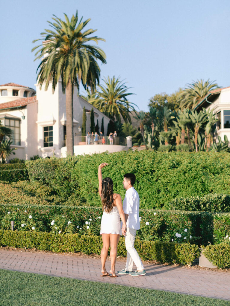 A couple walks hand in hand on a sunny pathway, surrounded by lush greenery and palm trees.