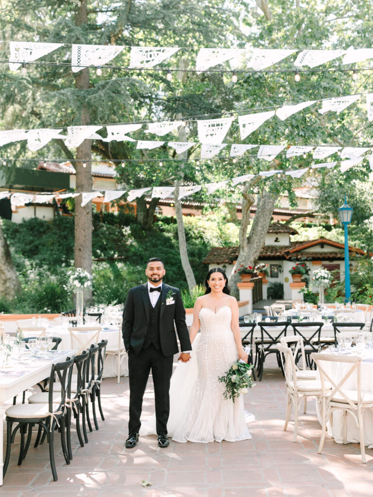 A smiling couple in wedding attire stands outdoors under decorative bunting. Surrounded by elegantly set table.