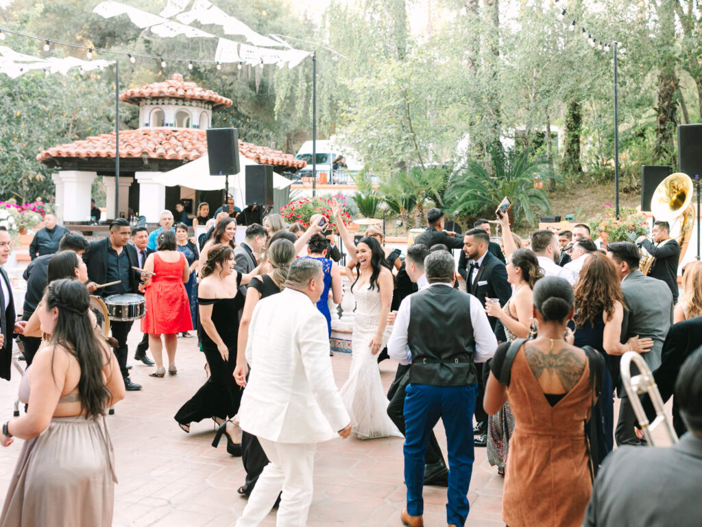 A vibrant outdoor wedding reception with a joyful bride dancing in the center, surrounded by guests in formal wear.