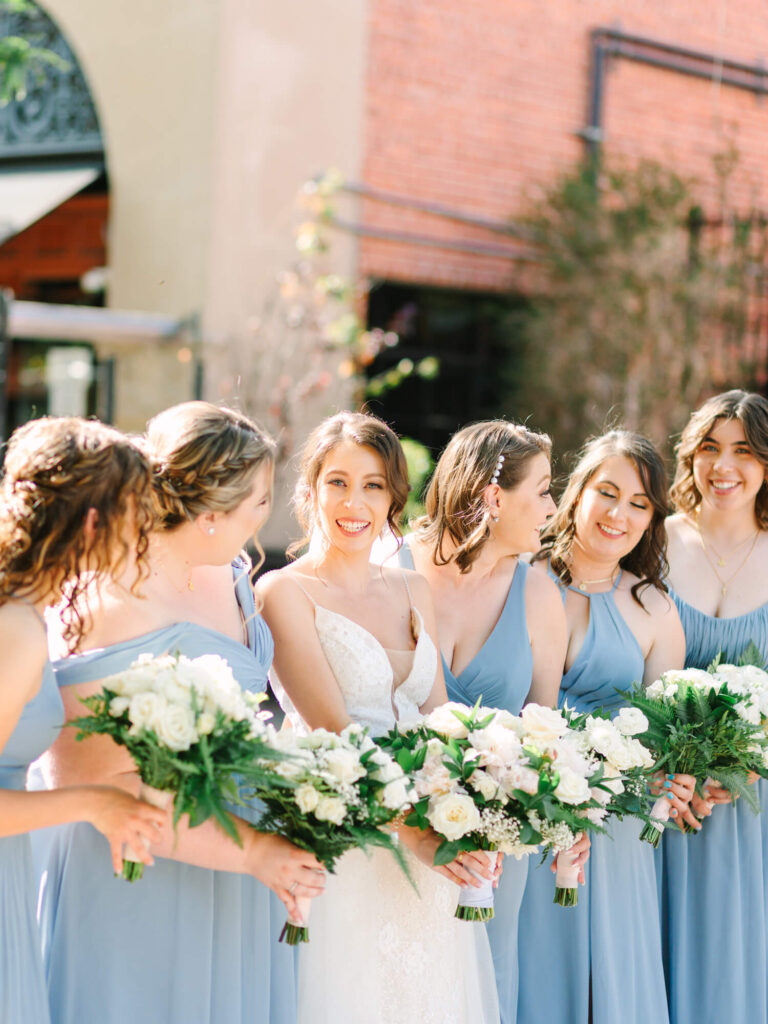 A smiling bride gazes at her groom in a brick-walled venue. Bridesmaids in the background hold bouquets.