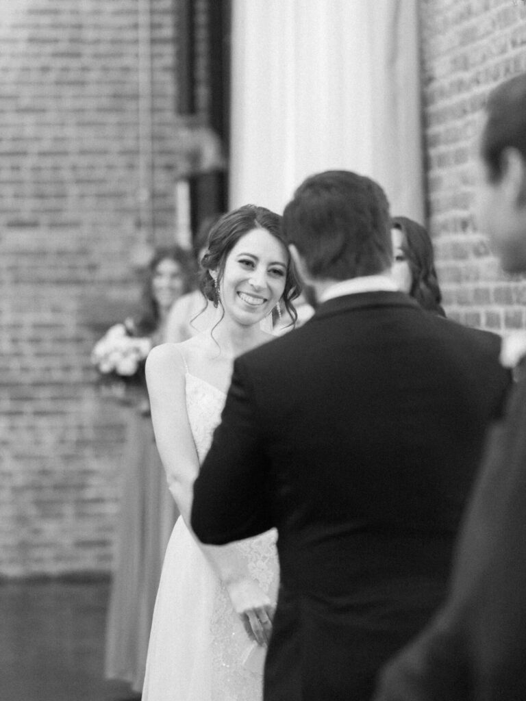 Bride in a white dress smiles warmly at a groom in a dark suit. Blurred bridesmaids in the background, with a brick wall beside them.