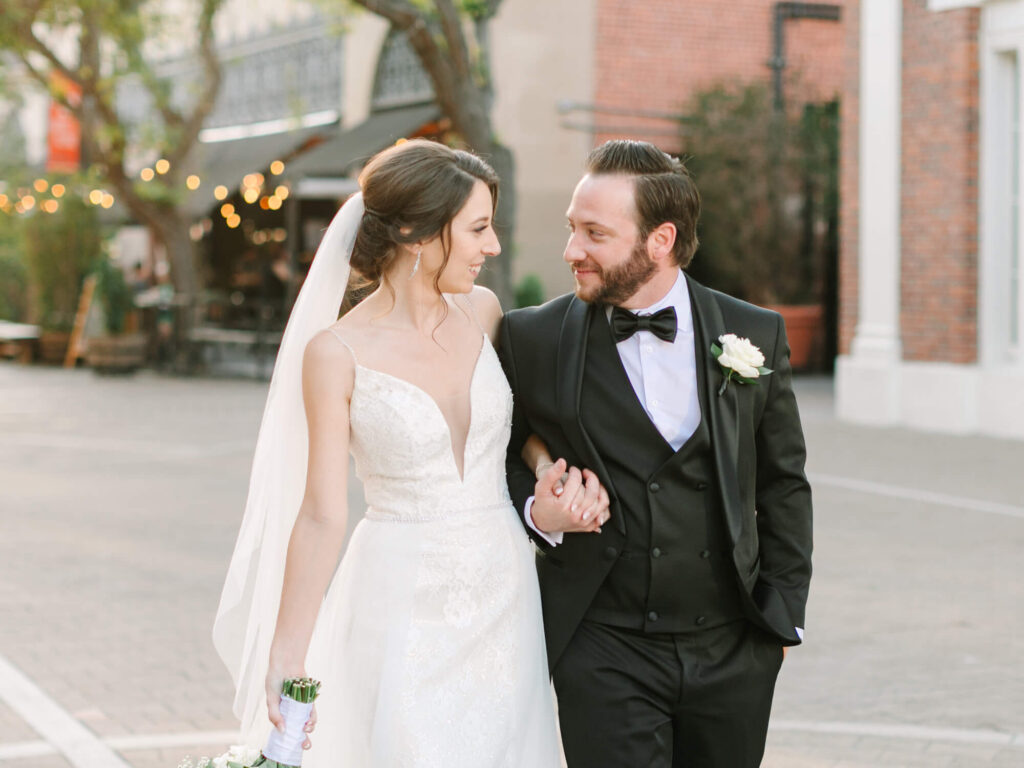 Bride and groom walk arm-in-arm on street, smiling warmly. Bride in lace gown and veil, groom in black tuxedo.