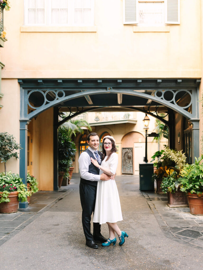 A couple embraces under an archway in a charming courtyard filled with potted plants and palm trees.
