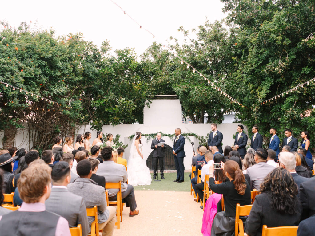 Outdoor wedding ceremony under string lights with a couple facing an officiant. Guests are seated on wooden chairs, surrounded by greenery.