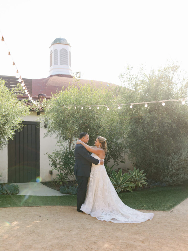 A couple dances joyfully outside under string lights and bright sun. The bride wears a flowing gown.