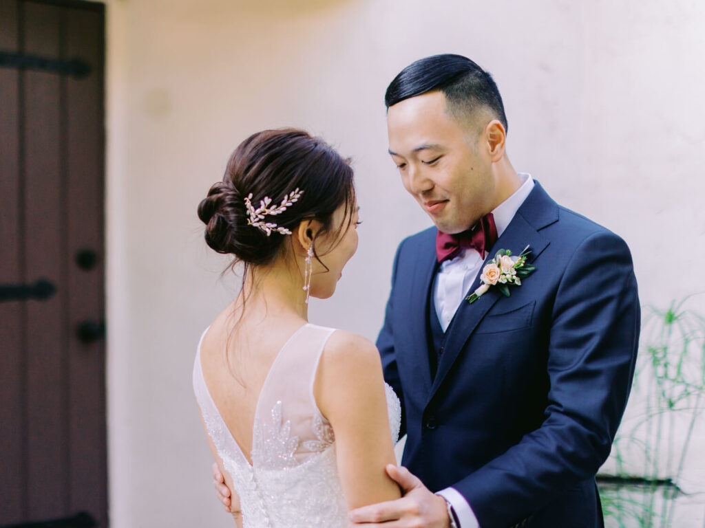 A bride and groom stand close, gazing at each other with joy. The bride wears a white gown and floral hairpiece, the groom a navy suit with a red bow tie.