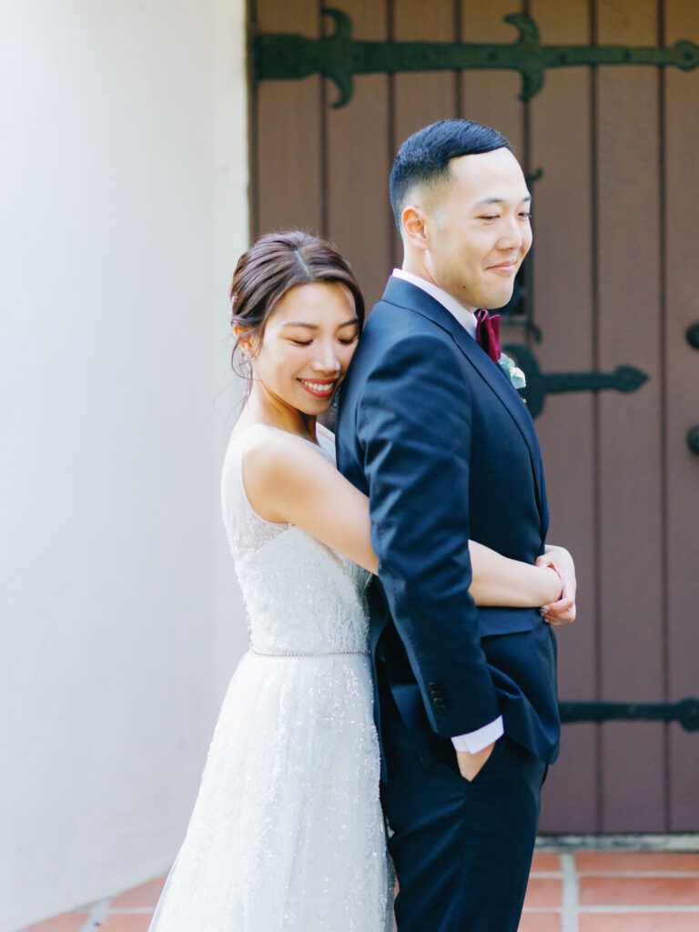 A bride in a white lace gown hugs a groom in a black suit from behind. They stand in front of a wooden door.