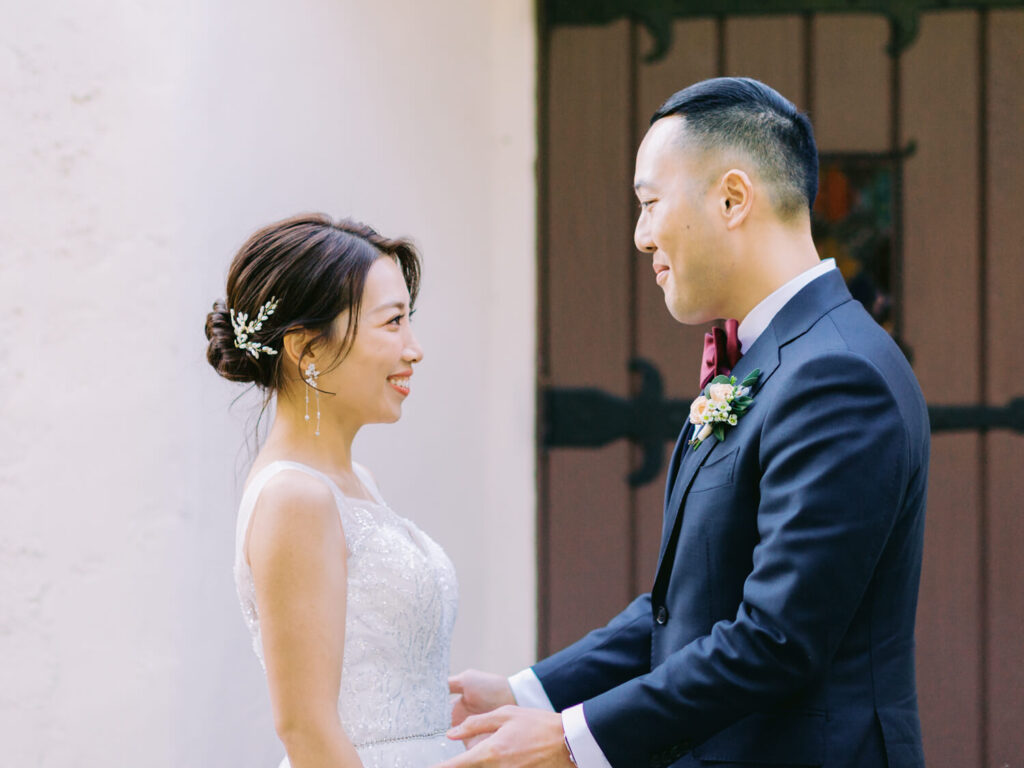 Bride and groom facing each other, smiling warmly. Bride in a white dress with floral hairpiece, groom in a black suit with a boutonnière.