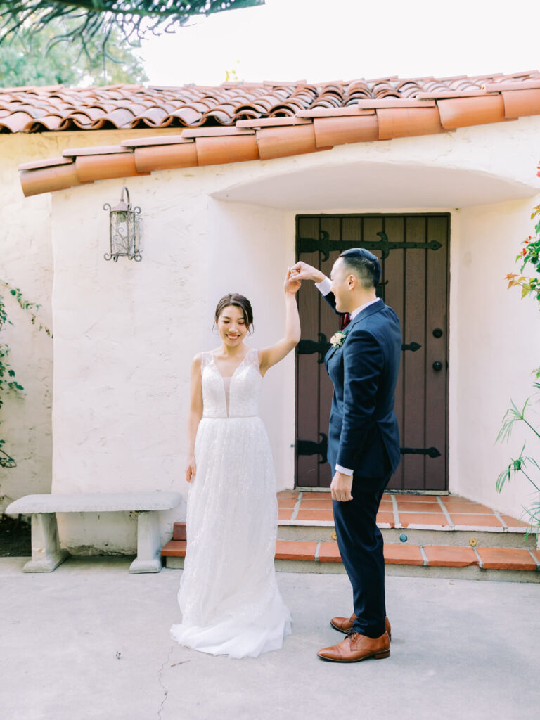 Bride in a white lace gown and groom in a dark suit share a joyful dance under a rustic archway with terracotta tiles.