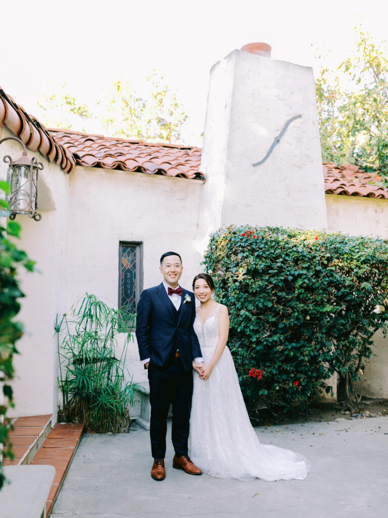 A couple in wedding attire smiles warmly, standing in front of a rustic white stucco building with a red-tiled roof, surrounded by greenery.