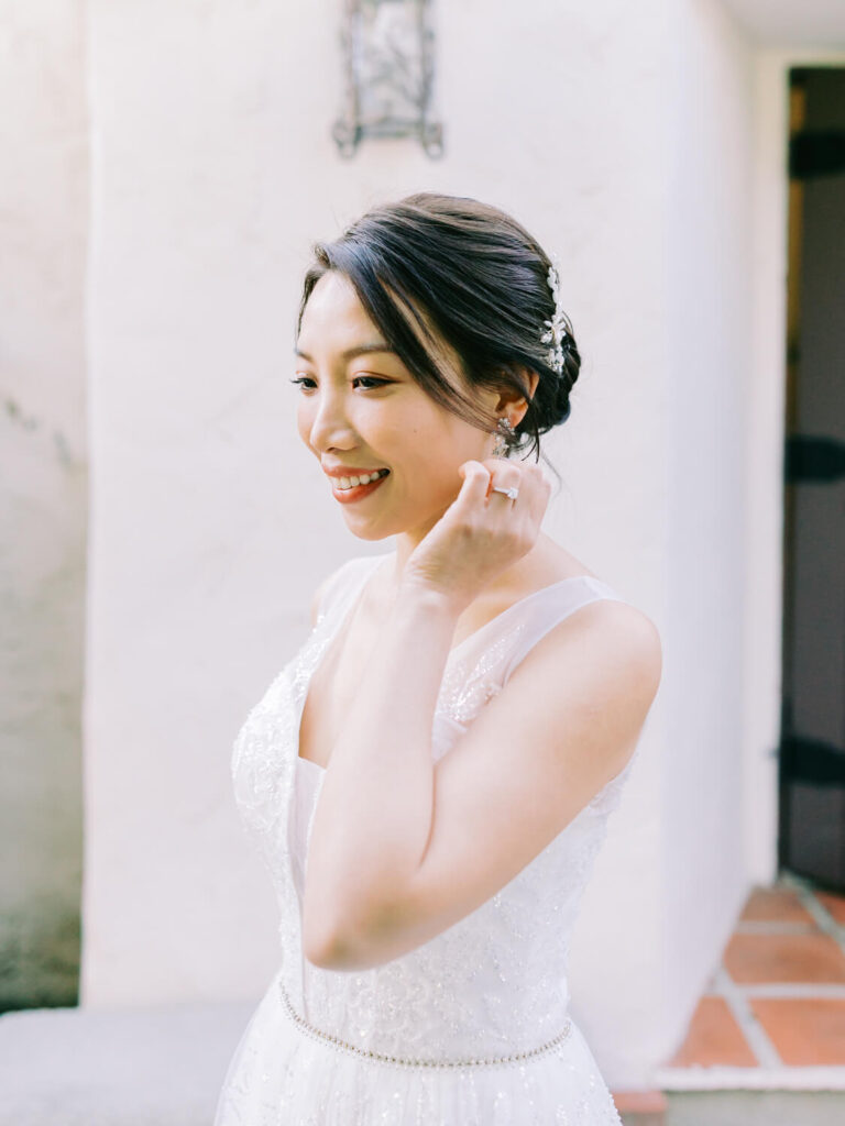 Bride in a white dress smiles joyfully, adjusting an earring. Her hair is elegantly styled with a decorative piece.