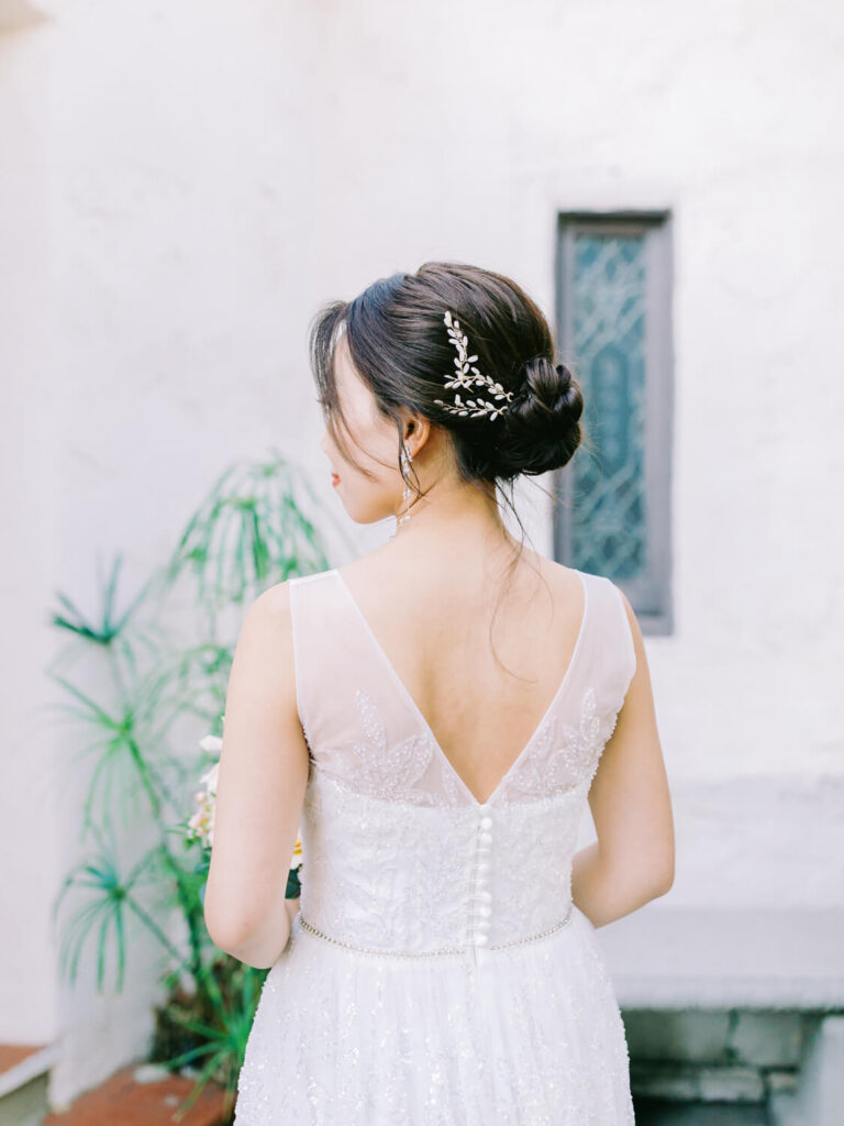 Bride with an elegant updo and hairpin in a lace wedding dress, back view. She stands in front of a textured wall.