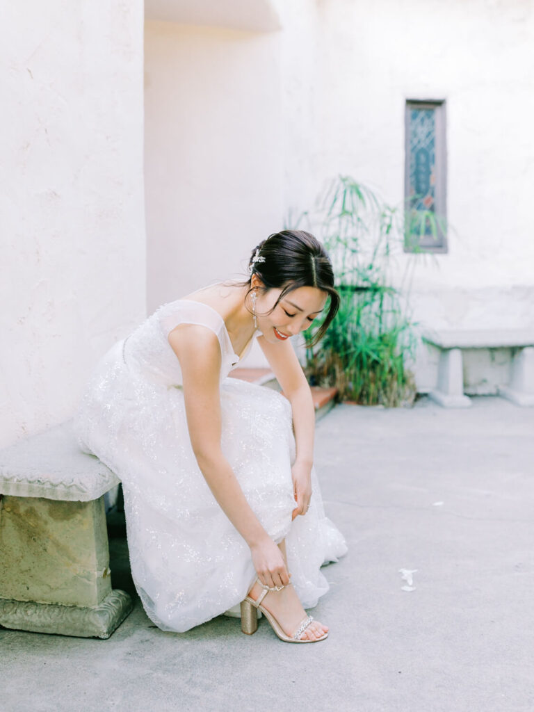 Bride sitting on a stone bench in a serene courtyard, adjusting her sparkling white gown and strappy heels, smiling softly, with plants nearby.