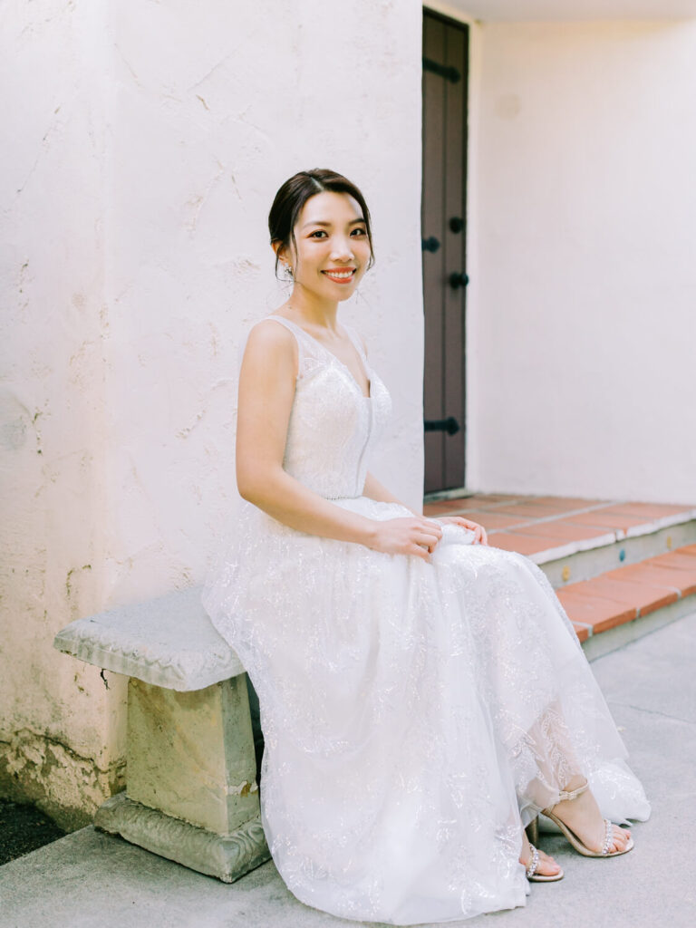 A woman in a white lace gown sits on a stone bench outside, smiling softly.