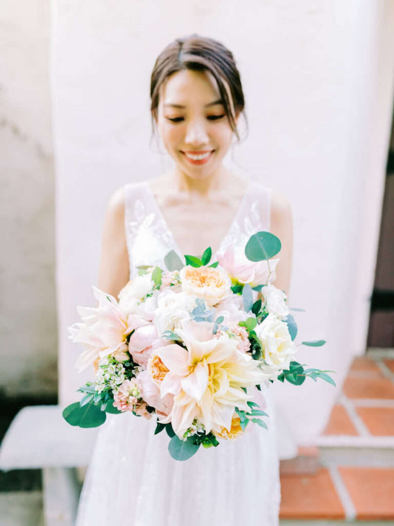 A bride with a joyful smile holds a vibrant bouquet of soft pink and white flowers, standing against a light, textured wall.