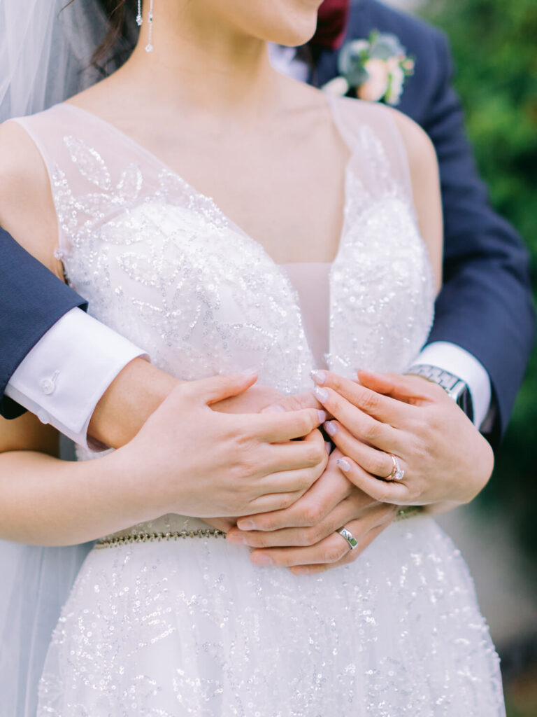 A bride in a sparkly white gown is embraced from behind by a groom in a dark suit. Their hands are interlocked, showing wedding rings.