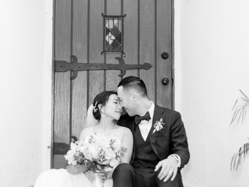 A bride and groom sit closely on a step, eyes closed, gently touching foreheads. The bride holds a bouquet, both are smiling softly.