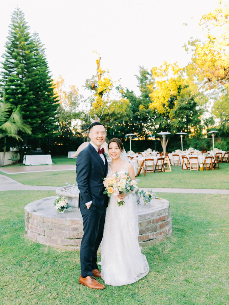 A bride and groom smile in front of a fountain surrounded by green grass at an outdoor wedding. Tables are set up in the background under warm, glowing lights.