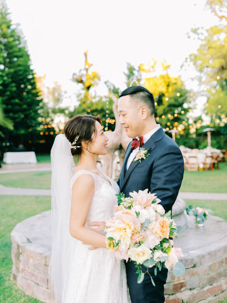 Bride in a white gown and groom in a navy suit stand by a fountain, smiling joyfully. They're surrounded by string lights and greenery, holding a bouquet.