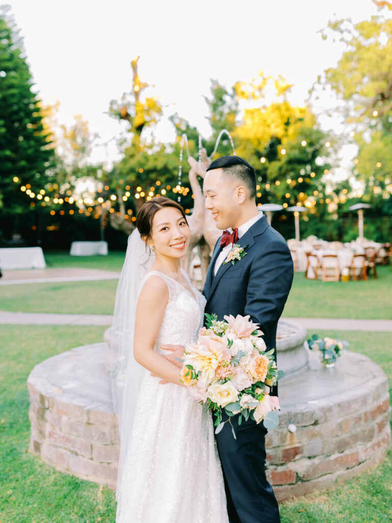 Bride in a white gown and groom in a navy suit stand by a fountain, smiling joyfully. They're surrounded by string lights and greenery, holding a bouquet.