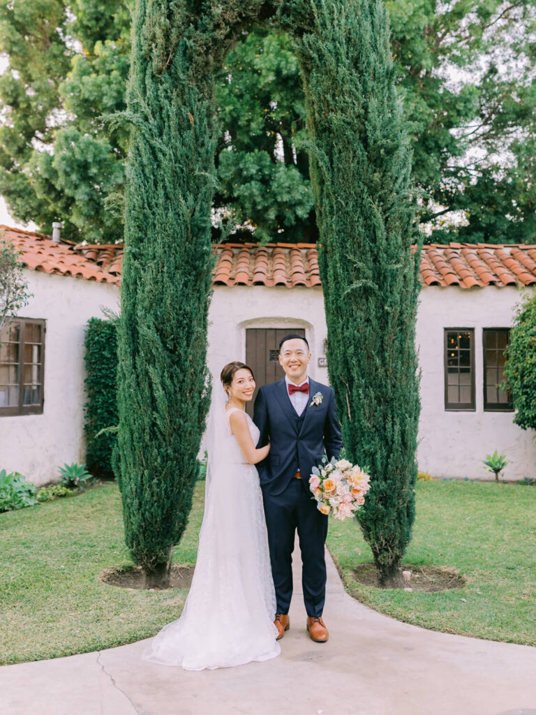 Bride in a white gown and groom in a navy suit and red bow tie stand smiling under an archway of tall trees, set in a lush garden.