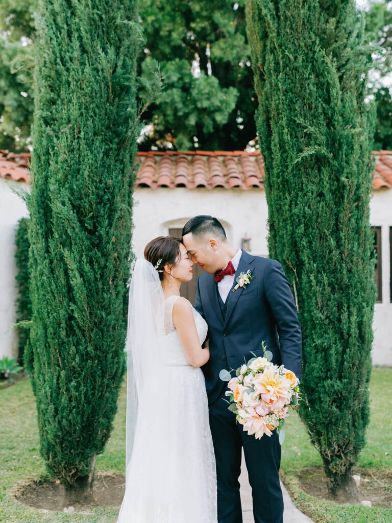 Bride and groom stand closely, foreheads touching, amid tall green trees.