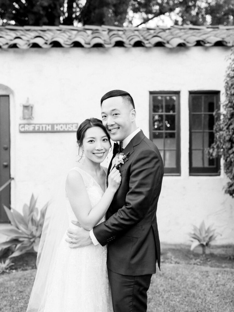 A joyful couple embraces in front of a building labeled "Griffith House." The bride wears a veil and gown; the groom is in a suit with a boutonniere.