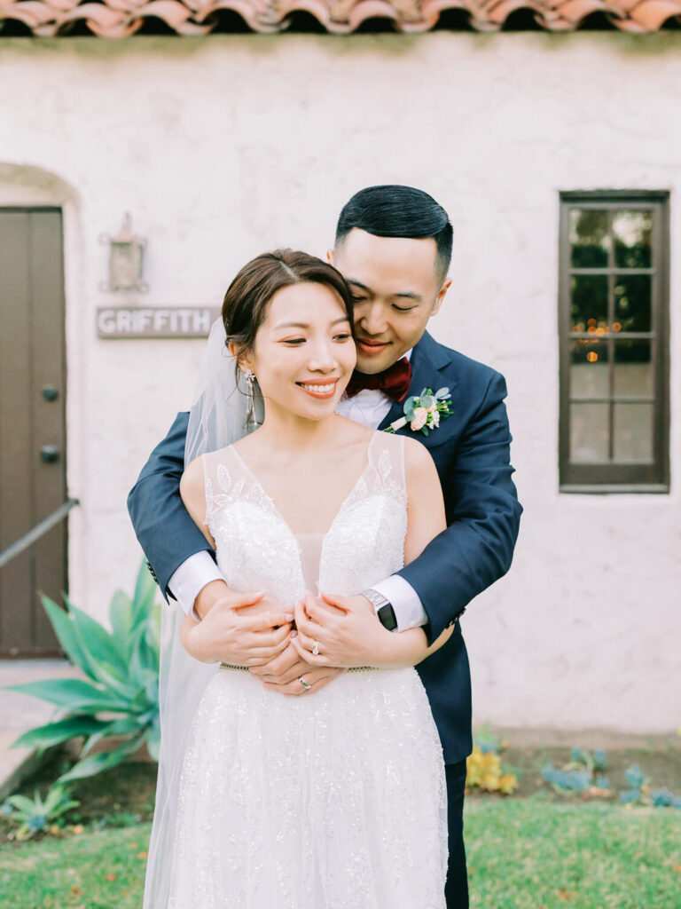 A bride and groom embrace joyfully in front of a rustic building labeled "Griffith House." The bride wears a white gown and veil; the groom, a navy suit.