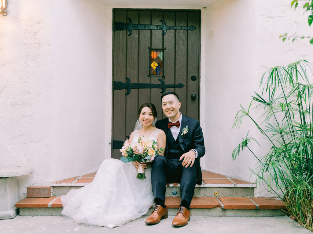 Bride in a white gown and groom in a navy suit sit on steps, smiling joyfully. She holds a bouquet, with a rustic wooden door in the background.