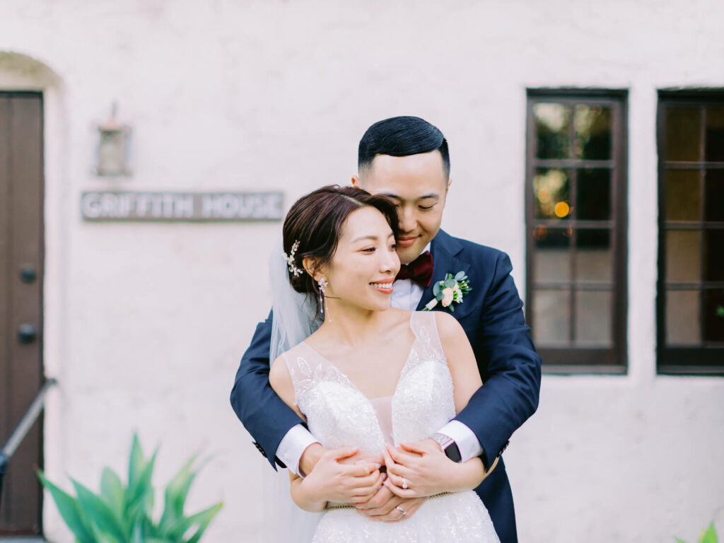A bride and groom embrace joyfully in front of a rustic building labeled "Griffith House." The bride wears a white gown and veil; the groom, a navy suit.