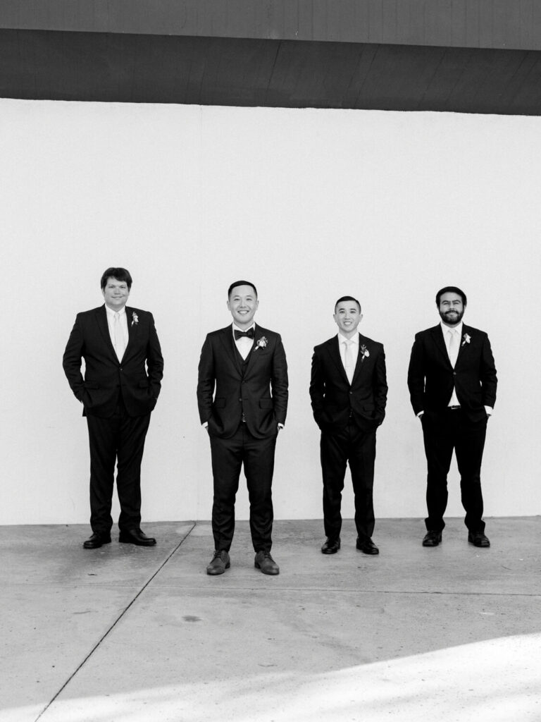 Four men dressed in suits stand in a line against a plain white wall. They appear cheerful, with hands in pockets, conveying a formal yet relaxed tone. Black and white photo.