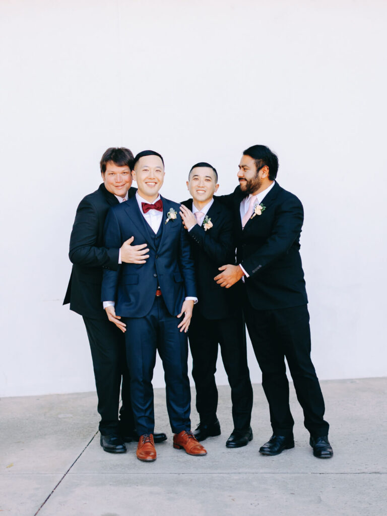 Four men in suits stand against a plain white background, smiling and laughing. The groom wears a red bow tie and brown shoes.