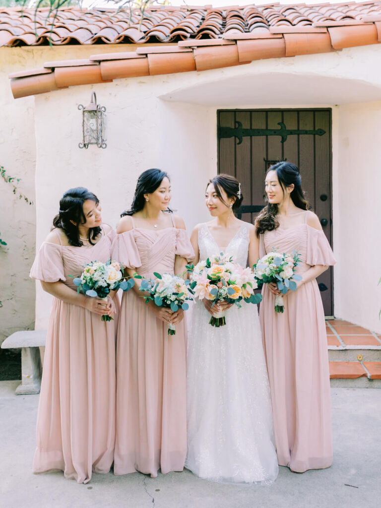 A bride in a white gown holds a bouquet, surrounded by three bridesmaids in blush pink dresses holding flowers. They're outside a building with a tiled roof, smiling warmly at each other.