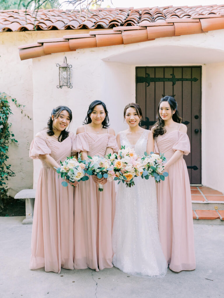 A bride in a white gown holds a bouquet, surrounded by three bridesmaids in blush pink dresses holding flowers. They're outside a building with a tiled roof.
