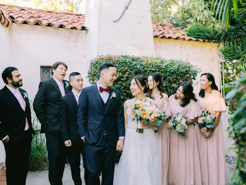 A joyful wedding party stands outdoors. The bride in a white dress holds a bouquet, smiles at the groom in a navy suit. Bridesmaids in pink dresses flank them, holding bouquets.