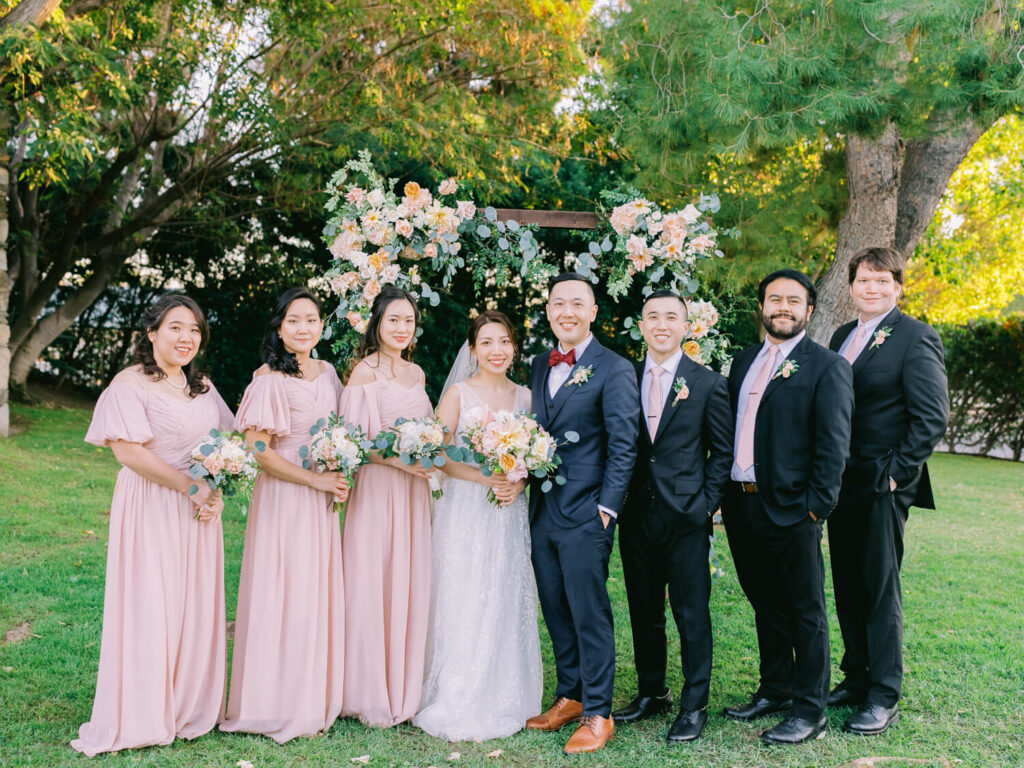 Bride in white with bridesmaids in pink dresses and groomsmen in black suits pose cheerfully under a floral arch in a verdant garden setting.